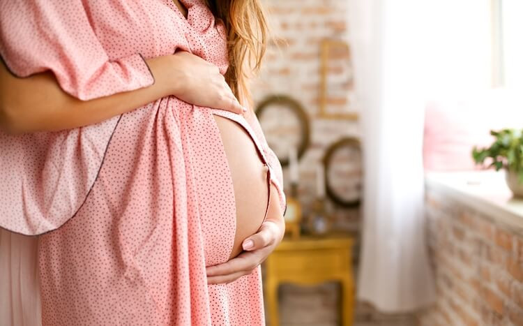 kobieta w ciąży czule obejmuje brzuch stojąc przy oknie